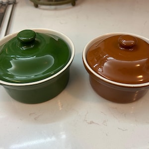 May include: Two ceramic ramekins with lids. One is a glossy forest green, the other is a rich brown. Both have white rims and small, round knobs on top. These small, lidded dishes are ideal for baking and serving individual portions.