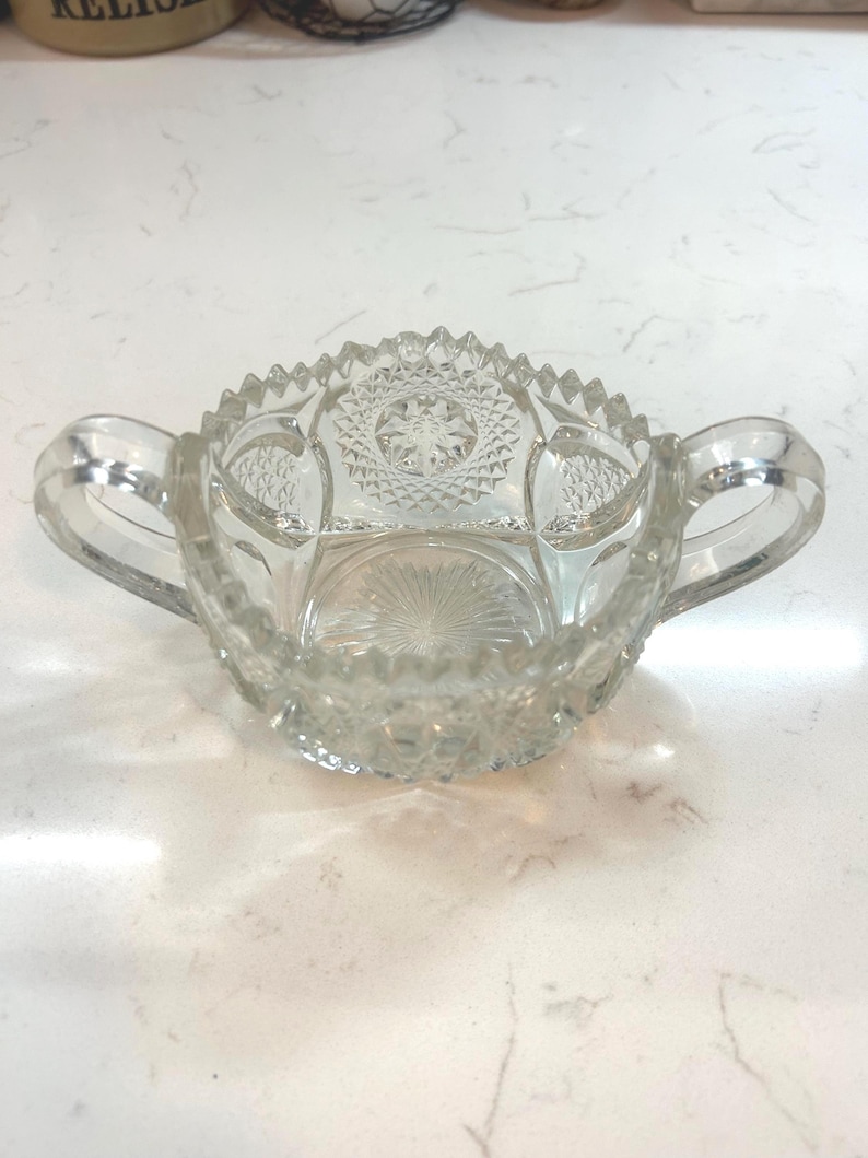 May include: Clear glass sugar bowl with two handles and a decorative, cut-glass pattern. The bowl has a scalloped rim and a starburst design on the base. A vintage piece for serving sugar or other condiments.