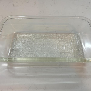 May include: A rectangular, clear glass baking dish with rounded corners and small handles on each side. The dish has a textured bottom and is designed for oven use. The glass is transparent, allowing visibility of the contents.