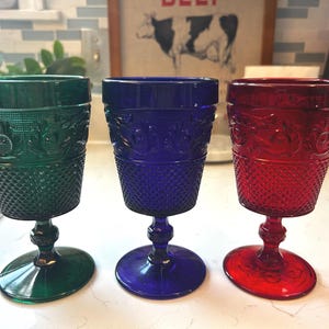 May include: Three glass goblets in emerald green, royal blue, and ruby red. Each goblet features a textured diamond pattern on the lower bowl and an embossed floral design around the upper rim, supported by a short, detailed pedestal stem.