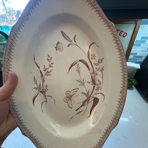 May include: Oval-shaped antique serving platter with a cream-colored base and a brown floral and butterfly design. The rim features a decorative brown border. A classic piece for serving food.