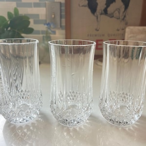 May include: Three clear, crystal drinking glasses with a diamond-cut pattern on the bottom. The glasses are tall and cylindrical, perfect for water or other beverages. The glasses are displayed on a white countertop.
