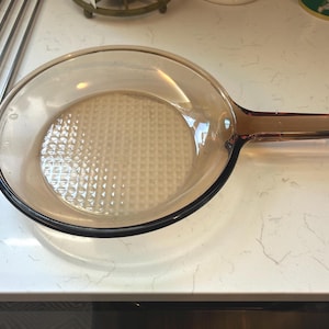 May include: A clear, amber-colored glass skillet with a long handle. The skillet has a textured bottom and a rounded shape. The handle is an extension of the skillet's body. The skillet is designed for cooking.