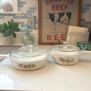 May include: Two white, handled casserole dishes with glass lids. Each dish features a colorful fruit and vegetable design and the words "La Ferme" on the side. The dishes are on a white countertop.
