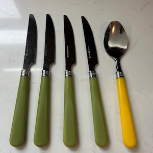Puede incluir: Un juego de cuatro cuchillos con mangos verde oliva y una cuchara con mango amarillo. Los cuchillos tienen hojas dentadas y detalles plateados. La cuchara tiene un cuenco plateado pulido. Los cubiertos están dispuestos sobre una superficie clara.
