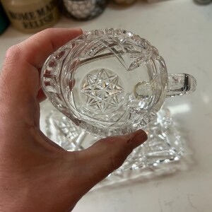 May include: A clear, cut-glass creamer with a handle, held in a hand. The creamer has a starburst design on the bottom and a geometric pattern on the sides. A matching rectangular dish is visible below.