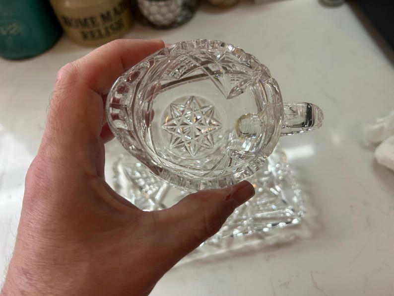 May include: A clear, cut-glass creamer with a handle, held in a hand. The creamer has a starburst design on the bottom and a geometric pattern on the sides. A matching rectangular dish is visible below.