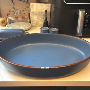 May include: A large, round, matte blue serving dish with a dark brown rim. The exterior features a smooth, solid blue finish, while the interior is a slightly lighter shade of blue. Small, light blue and white striped accents decorate the rim.