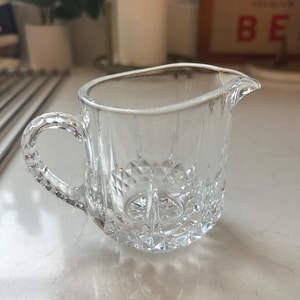 May include: A clear glass creamer with a handle and a spout. The creamer has a diamond-cut pattern on the bottom and vertical lines on the sides. The handle is also textured. The creamer is designed for serving liquids.