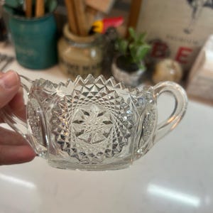 May include: Clear, vintage-style glass sugar bowl with two handles. The bowl features a starburst pattern on the base and sides, with a zig-zag design along the rim. The sugar bowl is held by a hand.