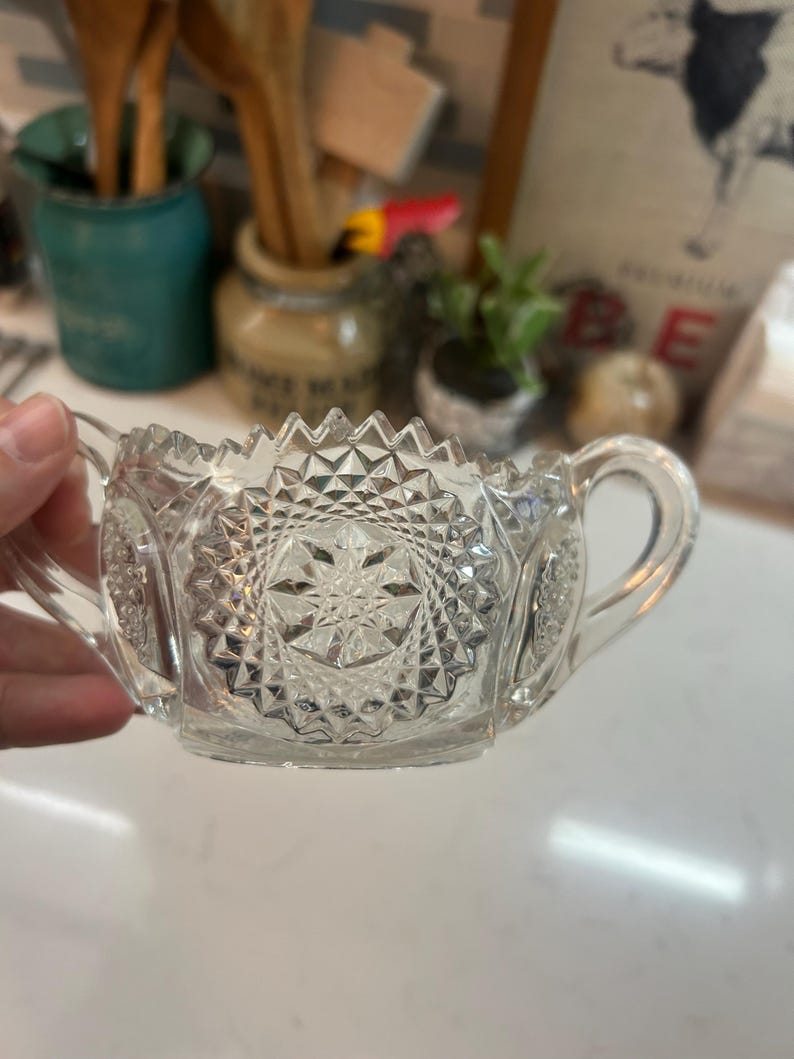 May include: Clear, vintage-style glass sugar bowl with two handles. The bowl features a starburst pattern on the base and sides, with a zig-zag design along the rim. The sugar bowl is held by a hand.