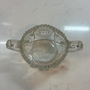 May include: Clear glass sugar bowl with two handles. The bowl has a decorative, textured pattern around the rim and on the base. The sugar bowl is sitting on a white surface.