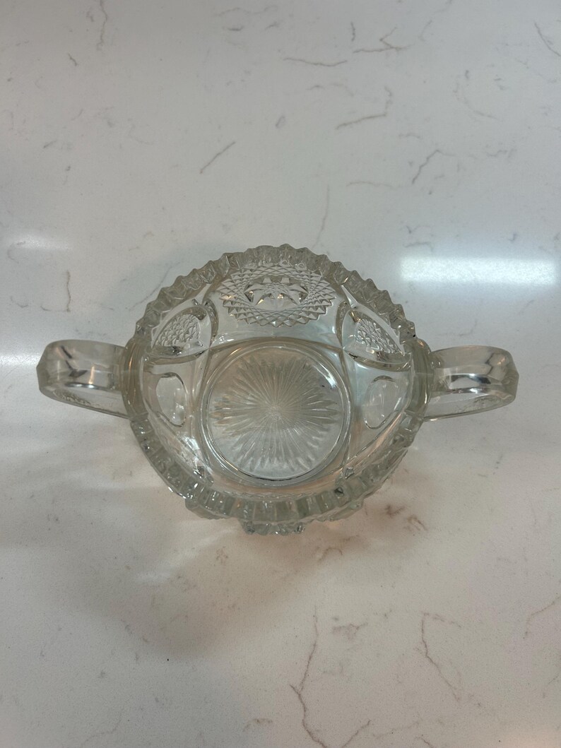 May include: Clear glass sugar bowl with two handles. The bowl has a decorative, textured pattern around the rim and on the base. The sugar bowl is sitting on a white surface.