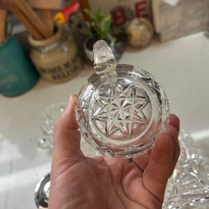 May include: A clear, round, cut-glass jar with a starburst pattern on the bottom. The jar has a small handle on top and is held in a person's hand. The jar is likely for storing condiments or small items.