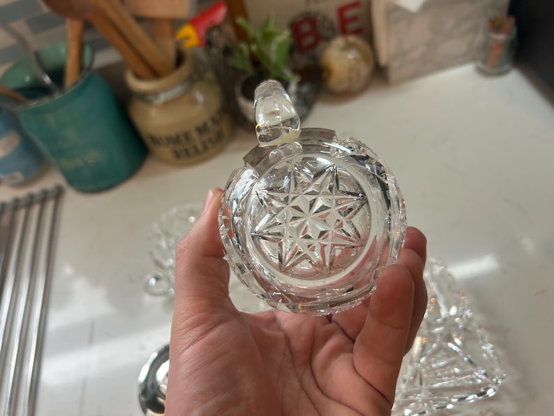 May include: A clear, round, cut-glass jar with a starburst pattern on the bottom. The jar has a small handle on top and is held in a person's hand. The jar is likely for storing condiments or small items.
