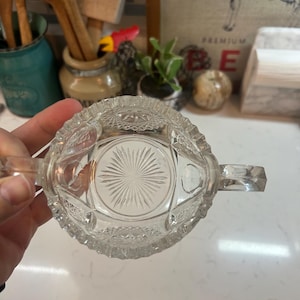 May include: Clear glass sugar bowl with two handles and a decorative, starburst pattern on the bottom. The bowl has a detailed, textured design around the rim and sides, adding to its vintage charm. A classic piece for serving sugar.