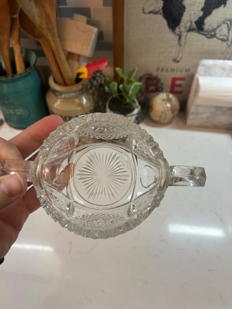 May include: Clear glass sugar bowl with two handles and a decorative, starburst pattern on the bottom. The bowl has a detailed, textured design around the rim and sides, adding to its vintage charm. A classic piece for serving sugar.