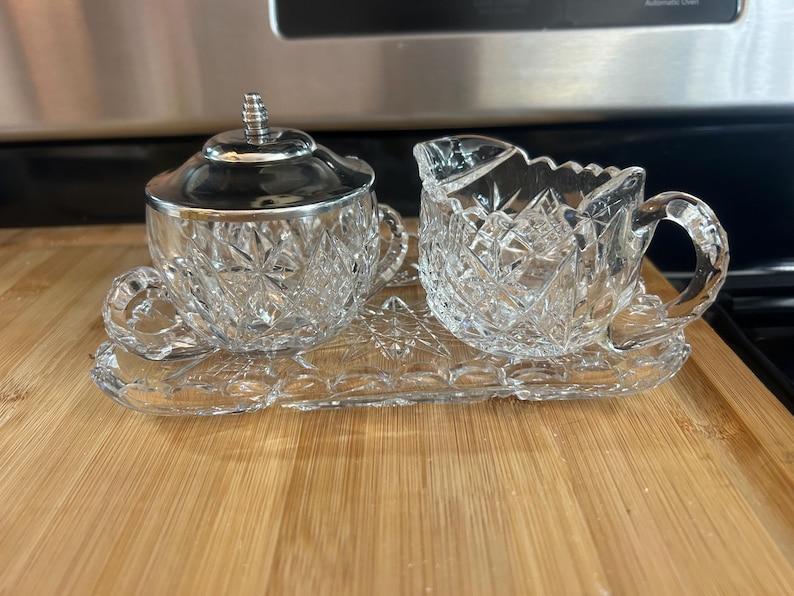 May include: A clear, cut-glass sugar bowl with a silver lid and a creamer sit on a matching tray. The sugar bowl and creamer have handles and are decorated with a starburst pattern. The tray is rectangular with a decorative edge.