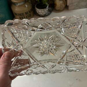 May include: A rectangular, clear glass dish with a starburst design in the center and star and geometric patterns. The edges have a scalloped design. This decorative dish is ideal for serving or display.