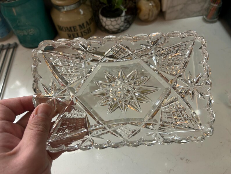 May include: A rectangular, clear glass dish with a starburst design in the center and star and geometric patterns. The edges have a scalloped design. This decorative dish is ideal for serving or display.