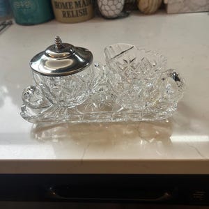 May include: A clear, cut-glass sugar and creamer set with a silver lid, resting on a matching tray. The set features a detailed, diamond-like pattern. The creamer has a handle, and the sugar bowl has a lid with a decorative finial.
