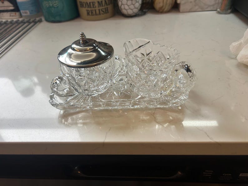 May include: A clear, cut-glass sugar and creamer set with a silver lid, resting on a matching tray. The set features a detailed, diamond-like pattern. The creamer has a handle, and the sugar bowl has a lid with a decorative finial.