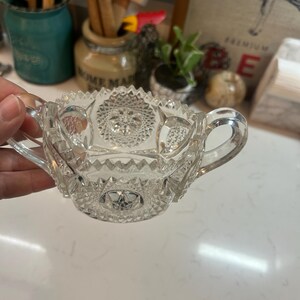 May include: Clear glass sugar bowl with two handles and intricate starburst and geometric patterns. The bowl has a scalloped rim and is likely vintage or antique. A classic piece for serving sugar or other condiments.