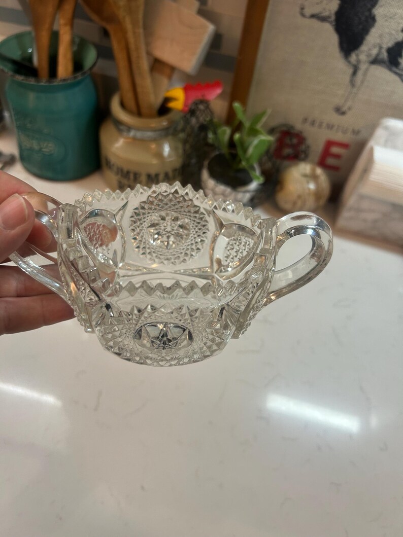 May include: Clear glass sugar bowl with two handles and intricate starburst and geometric patterns. The bowl has a scalloped rim and is likely vintage or antique. A classic piece for serving sugar or other condiments.