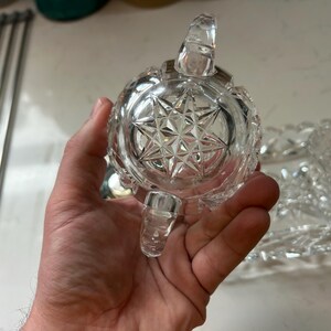 May include: A clear, cut-glass sugar bowl with a starburst pattern on the bottom. The bowl has a handle and a small spout. The bowl is held in a person's hand. A matching serving dish is in the background.
