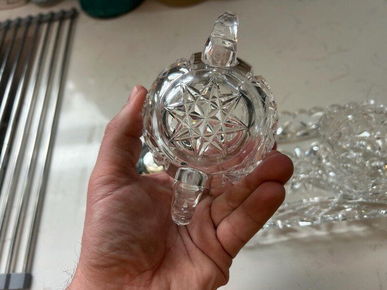 May include: A clear, cut-glass sugar bowl with a starburst pattern on the bottom. The bowl has a handle and a small spout. The bowl is held in a person's hand. A matching serving dish is in the background.