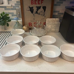 May include: A set of eight white ceramic ramekins arranged in a group on a countertop. Each ramekin features a smooth interior and a vertically ribbed exterior design. The bowls are uniform in size and color, presenting a clean, functional appearance.