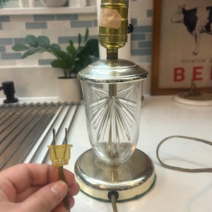 May include: A vintage table lamp with a clear, cut-glass shade and a brass base. The lamp has a decorative design on the glass and a standard light bulb socket. The electrical plug is visible. The lamp is on a white surface.