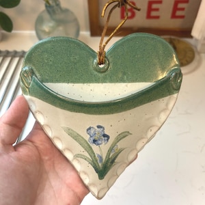 May include: Heart-shaped ceramic wall pocket with a green and white color scheme. The top is green, the middle is white, and the bottom is off-white with a floral design. A leather cord is attached for hanging.