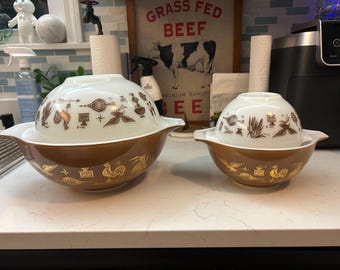 May include: Two vintage Pyrex mixing bowls. The larger bowl is brown with gold accents and a white lid with brown designs. The smaller bowl is nested inside. The bowls have a retro, mid-century modern aesthetic.
