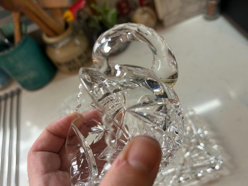 May include: A clear, cut-glass serving dish with a handle. The dish features a starburst and diamond pattern. The handle is curved and smooth. The dish is held in a hand.