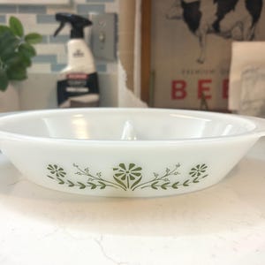 May include: White, round serving bowl with a floral design in olive green. The bowl has a slightly flared shape and two small handles. The floral pattern is symmetrical and repeats around the bowl's exterior.