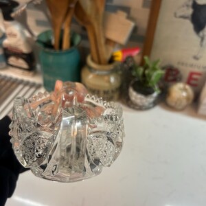 May include: A clear, cut-glass bowl with a handle, featuring intricate patterns and a scalloped rim. The bowl is held in a hand, with a blurred background of kitchen items.