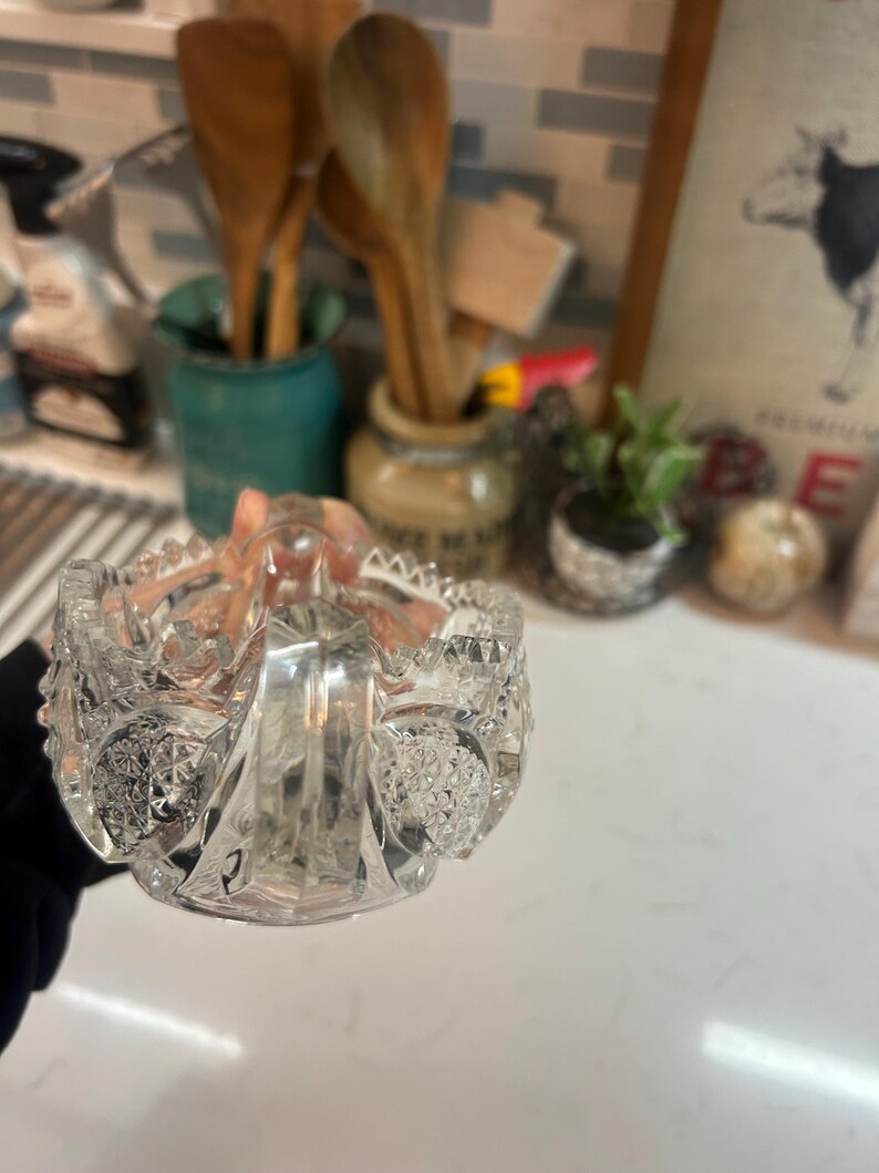 May include: A clear, cut-glass bowl with a handle, featuring intricate patterns and a scalloped rim. The bowl is held in a hand, with a blurred background of kitchen items.