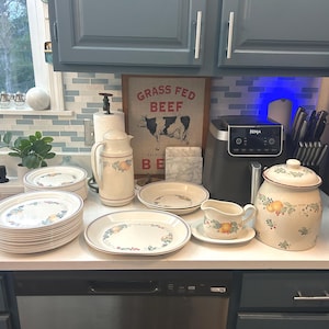 May include: A collection of cream-colored dinnerware with a floral design, including plates, bowls, and serving pieces. A matching pitcher and a large jar with a lid are also present. A sign that says "Grass Fed Beef" is visible in the background.