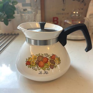 May include: A white teapot with a black handle and a metal lid. The body features a colorful botanical illustration of fruits and leaves with the text Le Thé printed in black script below the design. A silver metal band encircles the upper neck.