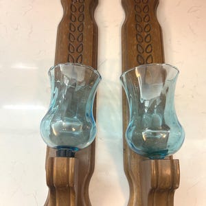 May include: Pair of vintage wall sconces. Each sconce features a wooden backplate with a decorative carved design and a clear blue glass candle holder. The sconces are designed to hold candles and add a touch of vintage charm to any room.