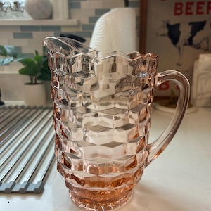 May include: A pink glass pitcher with a geometric diamond pattern. The pitcher has a handle and a spout for pouring. The pitcher is likely vintage and is sitting on a white countertop.