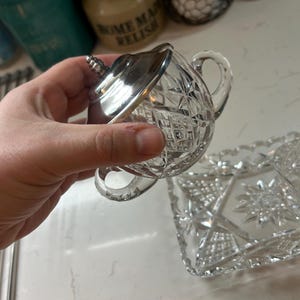 May include: A clear, cut-glass sugar bowl with a silver lid and handle. The bowl features a diamond pattern and a small handle. A matching rectangular serving tray is also visible.
