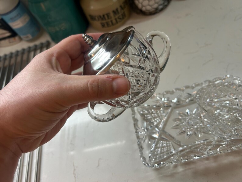 May include: A clear, cut-glass sugar bowl with a silver lid and handle. The bowl features a diamond pattern and a small handle. A matching rectangular serving tray is also visible.
