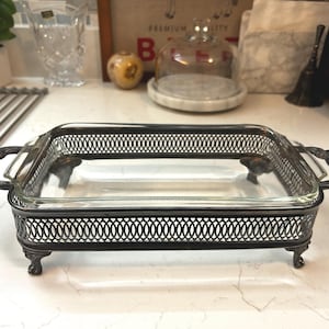 May include: A rectangular clear glass serving dish set within a silver-toned metal frame. The frame features an intricate lattice pattern on the sides, ornate handles on both ends, and four decorative feet that elevate the dish from the surface.