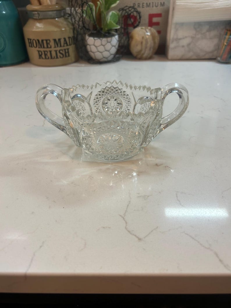 May include: A clear, cut-glass sugar bowl with two handles. The bowl has a detailed, starburst pattern on the bottom and around the rim. The handles are curved and attached to the sides of the bowl. A classic piece for serving sugar or other condiments.