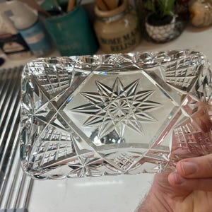 May include: A rectangular, clear crystal dish with a starburst design in the center. The dish has a geometric pattern of diamond shapes and is held in a hand. The dish is likely used for decorative purposes.