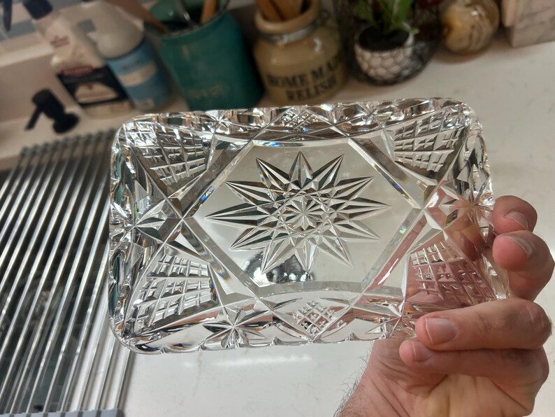 May include: A rectangular, clear crystal dish with a starburst design in the center. The dish has a geometric pattern of diamond shapes and is held in a hand. The dish is likely used for decorative purposes.