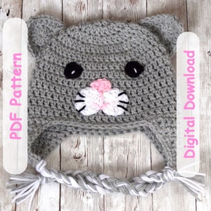 May include: Gray crocheted cat hat with white and pink details. The hat has a braided tail with white and gray yarn. The text "PDF Pattern" and "Digital Download" is on the left and right sides of the image.