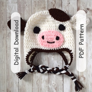 May include: A handmade crochet cow hat in white with brown spots and ears, a pink muzzle, and black eyes. The hat has a braided chin strap with brown and white strands. Text on the side reads "Digital Download" and "PDF Pattern".